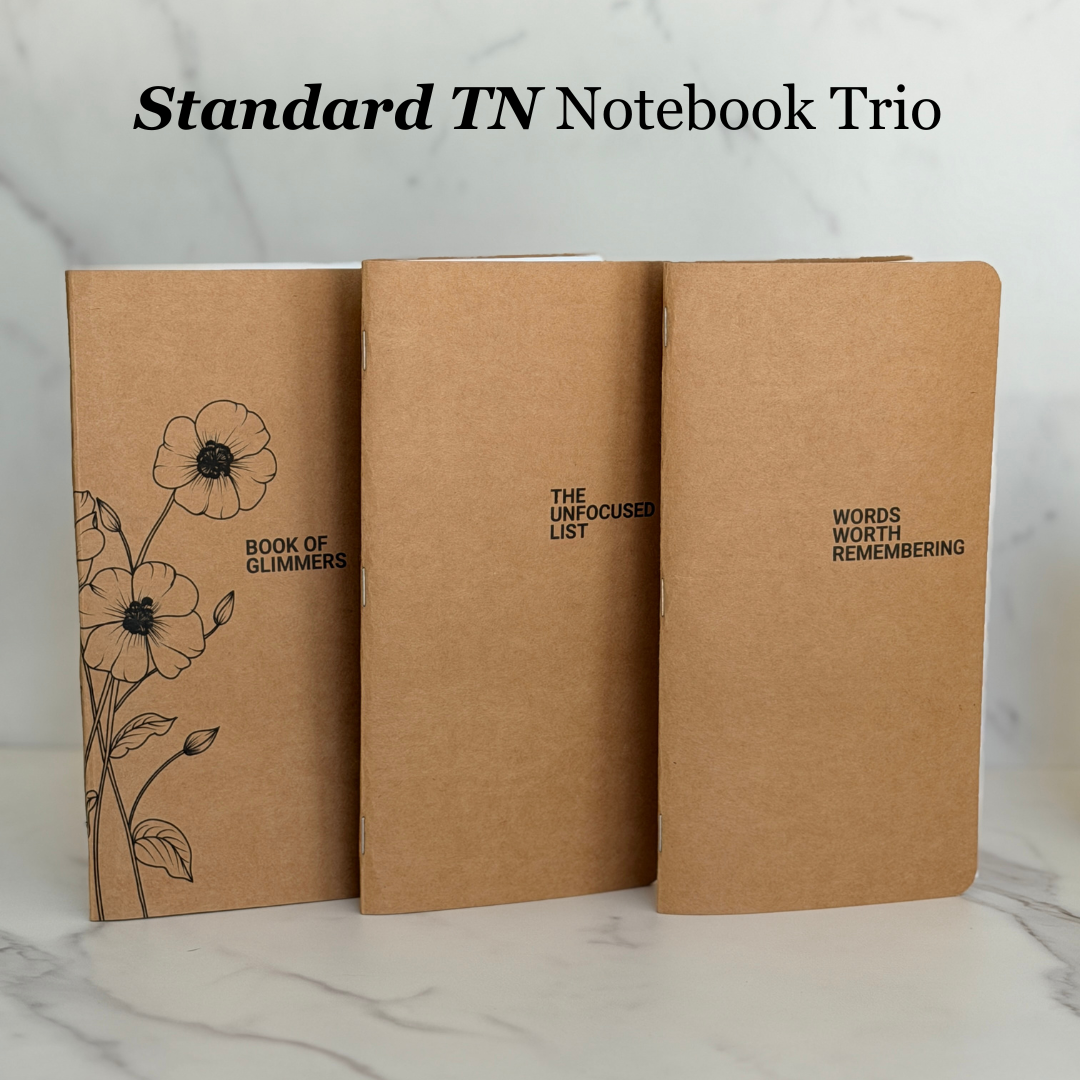 Three tan kraft-paper notebooks standing upright on a light marble surface, with the title 'Standard TN Notebook Trio' above them. The left notebook has black floral line art and the text 'Book of Glimmers'; the center says 'The Unfocused List'; the right says 'Words Worth Remembering'.