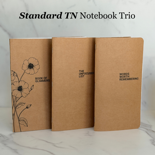 Three tan kraft-paper notebooks standing upright on a light marble surface, with the title 'Standard TN Notebook Trio' above them. The left notebook has black floral line art and the text 'Book of Glimmers'; the center says 'The Unfocused List'; the right says 'Words Worth Remembering'.